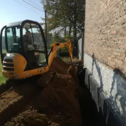 Żółta mini koparka JCB wykonuje wykop wzdłuż fundamentów ceglanego budynku, widoczna czarna folia ochronna na ścianie fundamentowej.