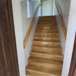 Widok z dołu na drewniane schody z jasnymi poręczami, prowadzące na górę przez białą futrynę drzwiową, z widocznym fragmentem brązowych drzwi po lewej stronie i wykończeniem ścian w jasnych kolorach.