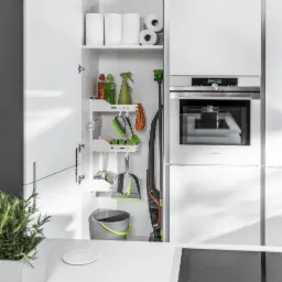 Wnętrze nowoczesnej, białej kuchni z otwartą szafką, w której znajdują się środki czystości, szczotki, zmiotka, szufelka, odkurzacz pionowy i rolki papieru, obok zabudowany piekarnik Siemens.