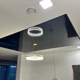 PIOFACH SPÓŁKA Z OGRANICZONĄ ODPOWIEDZIALNOŚCIĄ - Czarny, błyszczący sufit podwieszany z dwiema nowoczesnymi lampami pierścieniowymi, zabezpieczone folią fragmenty ścian w trakcie prac wykończeniowych.