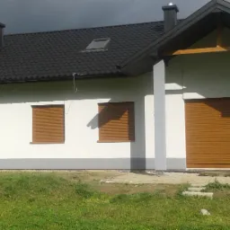 Biały dom z szarą podmurówką, brązowymi roletami zewnętrznymi i ciemnym dachem pod pochmurnym niebem.