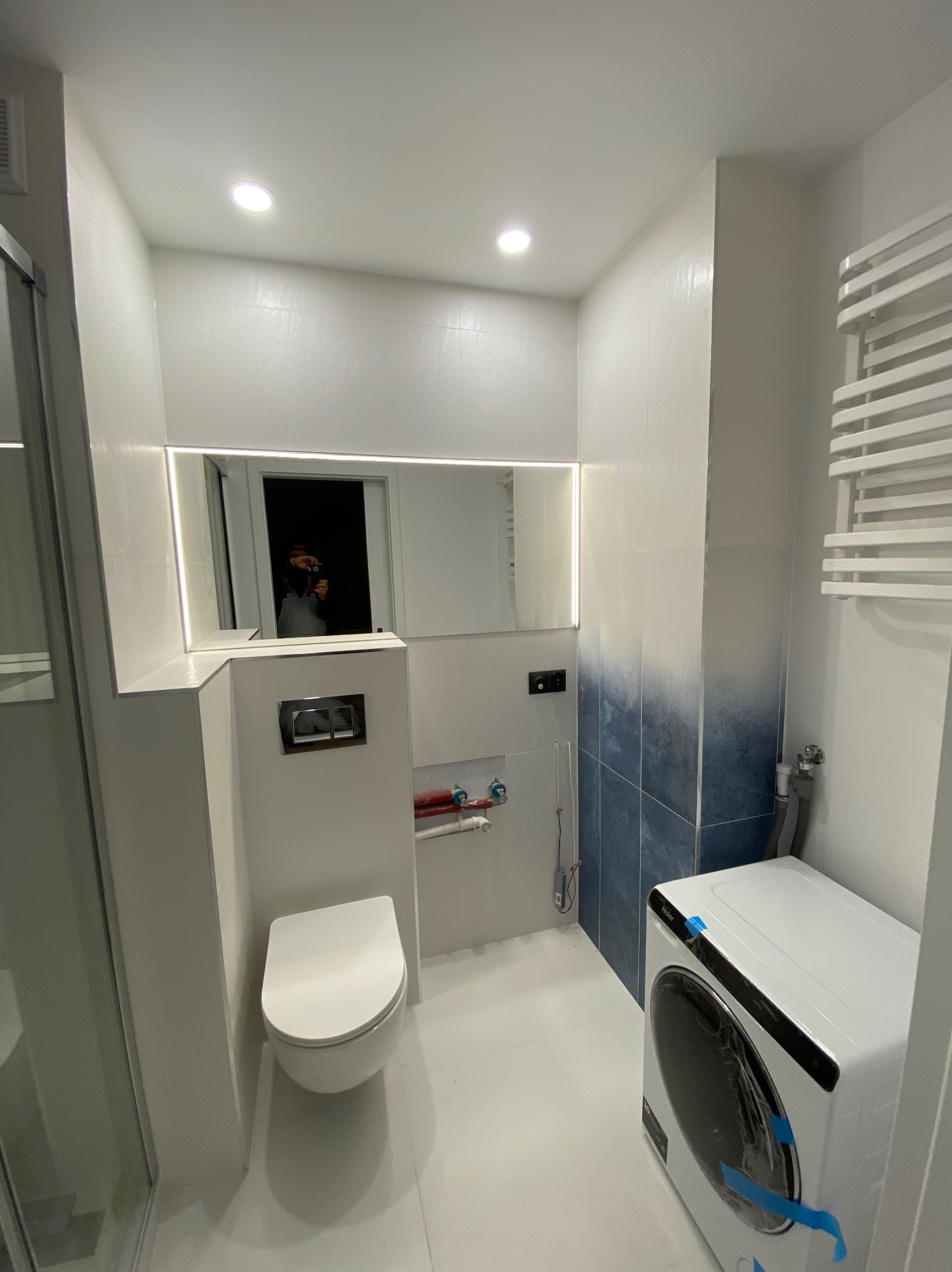 Nowoczesna łazienka z białymi i niebieskimi płytkami ombre, podświetlanym lustrem, podwieszaną toaletą i pralką w folii ochronnej.