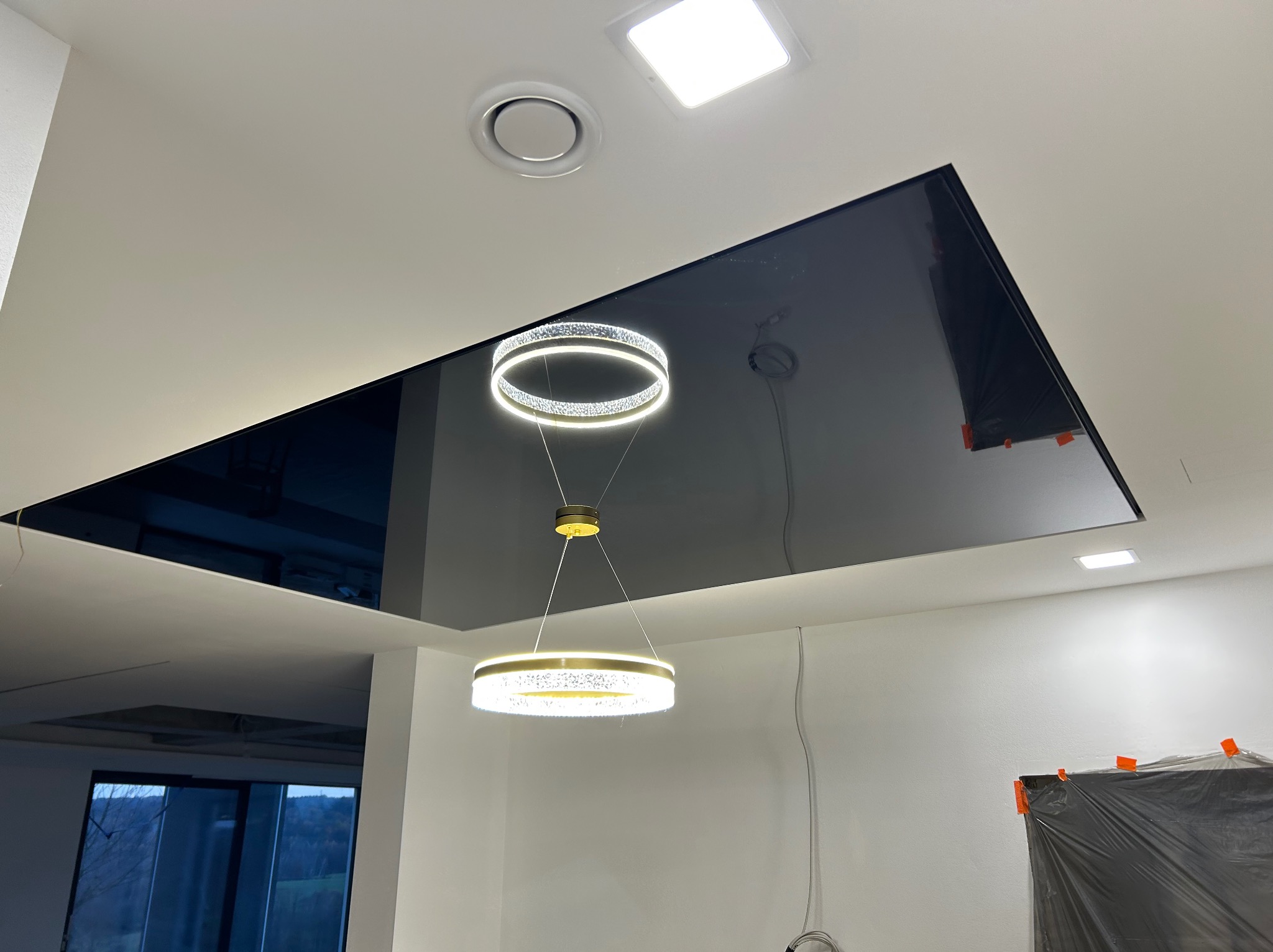 Czarny, błyszczący sufit podwieszany z dwiema nowoczesnymi lampami pierścieniowymi, zabezpieczone folią fragmenty ścian w trakcie prac wykończeniowych.