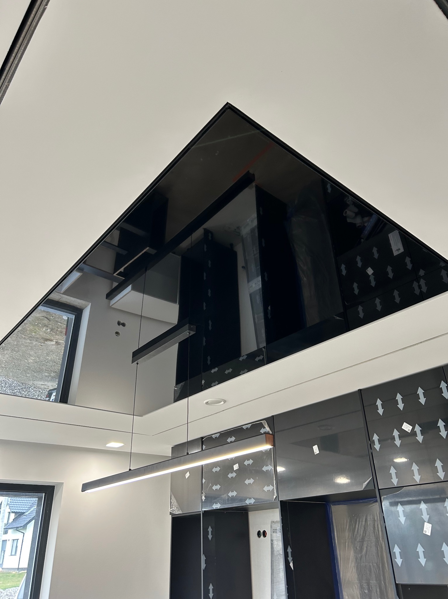 Nowoczesne wykończenie sufitu z czarnym, lustrzanym panelem w kształcie trójkąta, odbijającym elementy wnętrza, widoczna minimalistyczna lampa i częściowo zakryte folią ochronną czarne szafki...