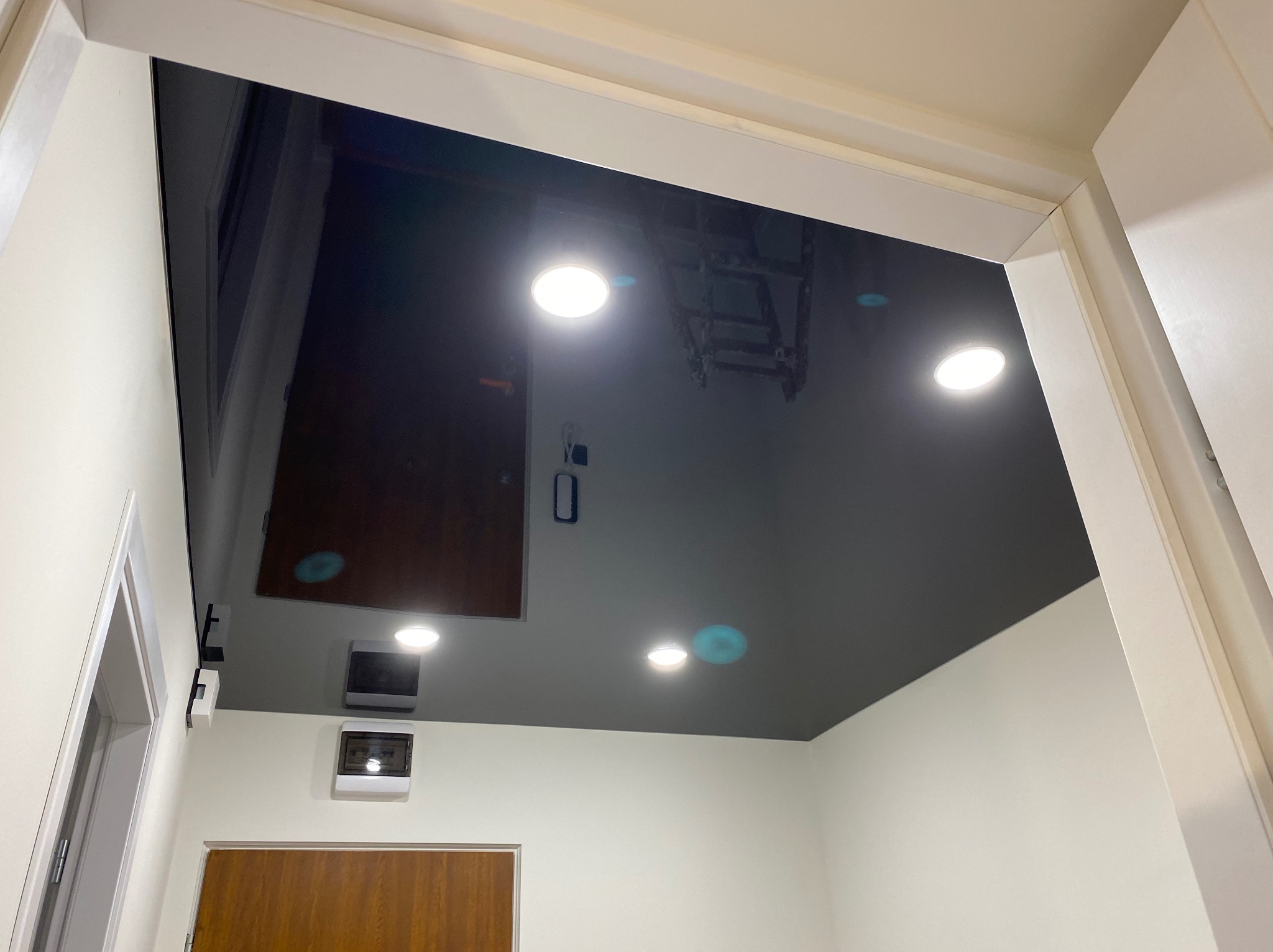 Błyszczący, czarny sufit napinany z wbudowanym oświetleniem punktowym, widziany z dołu, z widocznymi elementami instalacji i odbiciem drabiny.