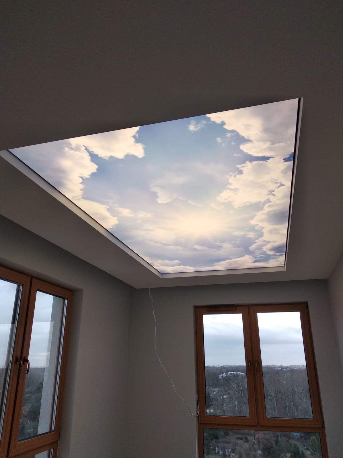 Pokój z podświetlanym sufitem imitującym niebo z chmurami, widoczne dwa okna z drewnianą ramą i szarymi ścianami.
