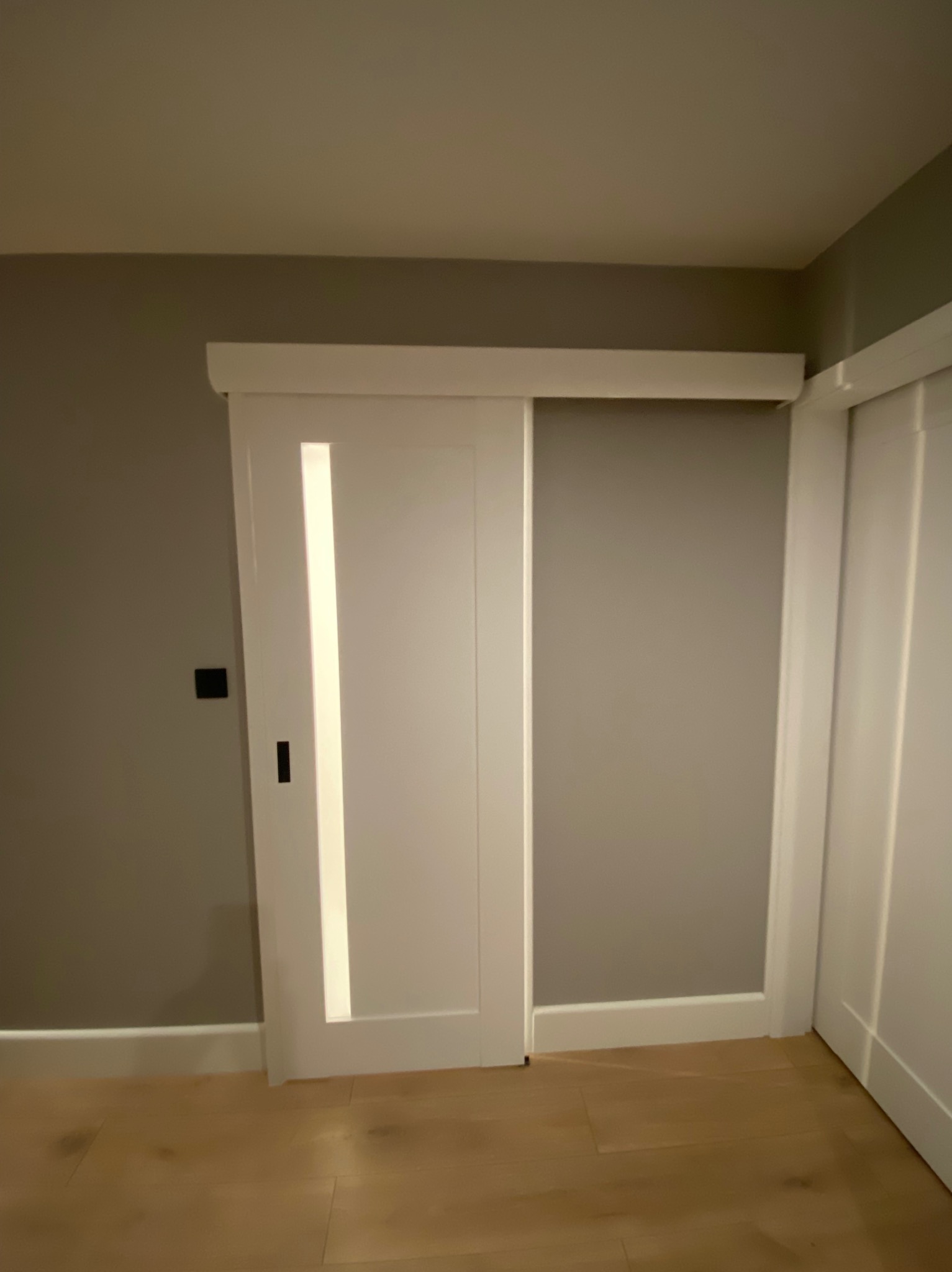 Białe, przesuwne drzwi z podłużnym, oświetlonym panelem w szarej ścianie, widoczny fragment drewnianej podłogi i czarny włącznik światła.