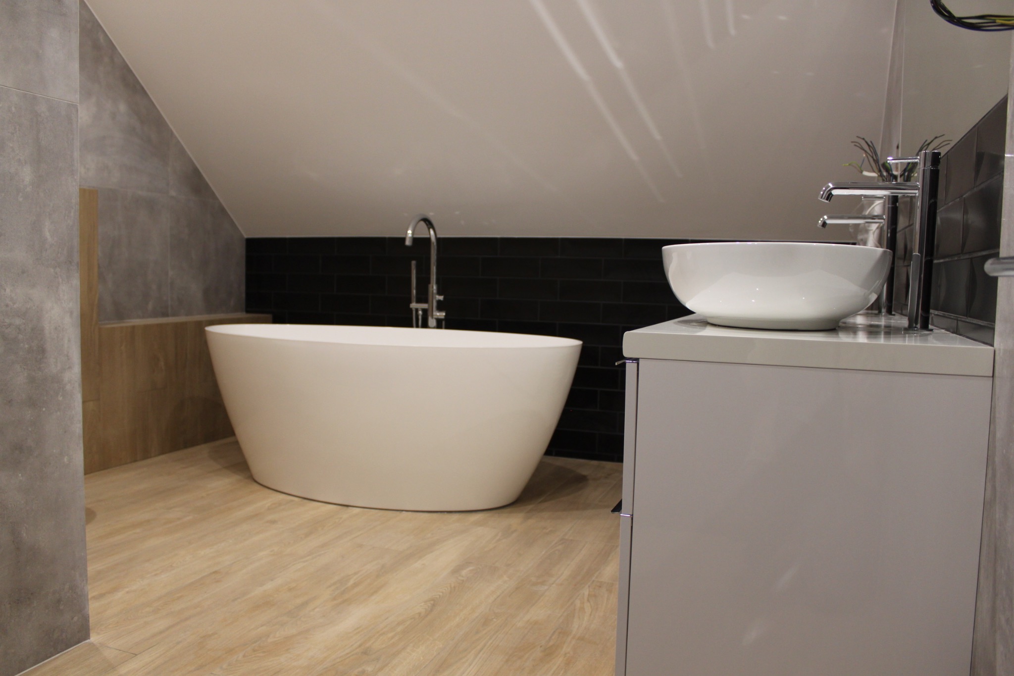 Nowoczesna łazienka na poddaszu ze skośnym sufitem, z wolnostojącą białą wanną, umywalką nablatową na szarej szafce i czarnymi płytkami na ścianie.