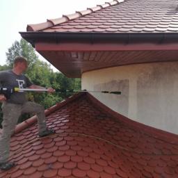 ARTEON - Mężczyzna w szarej koszulce i spodniach khaki, stojący na dachu krytym czerwoną dachówką, czyści elewację budynku za pomocą myjki ciśnieniowej z długą lancą. Widoczne ślady czyszczenia na jasnej...