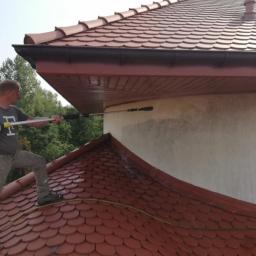 ARTEON - Mężczyzna w szarej koszulce i spodniach, stojąc na czerwonym dachu z dachówki, czyści jasną elewację domu za pomocą myjki ciśnieniowej z długą lancą. Widoczne ślady czyszczenia na elewacji.
