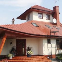 ARTEON - Pracownik w kombinezonie ciśnieniowo czyści czerwoną dachówkę domu jednorodzinnego z drabiny, widoczne zacieki i odbarwienia na dachu.