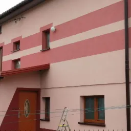 Dwukondygnacyjny dom z elewacją w odcieniach różu i beżu, z poziomymi pasami, brązowymi oknami i drzwiami. Na pierwszym planie drabina oparta o ścianę i sznur na pranie z przypiętymi klamerkami.