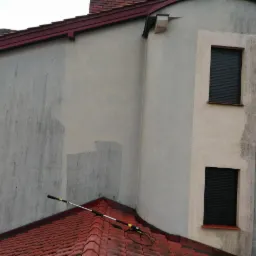 Fragment elewacji budynku z widocznymi śladami czyszczenia, gdzie kontrastuje obszar umyty z brudną powierzchnią; na pierwszym planie widoczny element roboczy lancy teleskopowej na tle czerwonej...