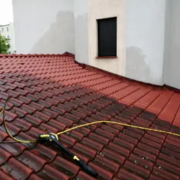 Częściowo oczyszczony dach z czerwonej dachówki, widoczna różnica w kolorze po umyciu, dysza myjąca ciśnieniowa z żółtym wężem na pierwszym planie, fragment elewacji budynku w tle.