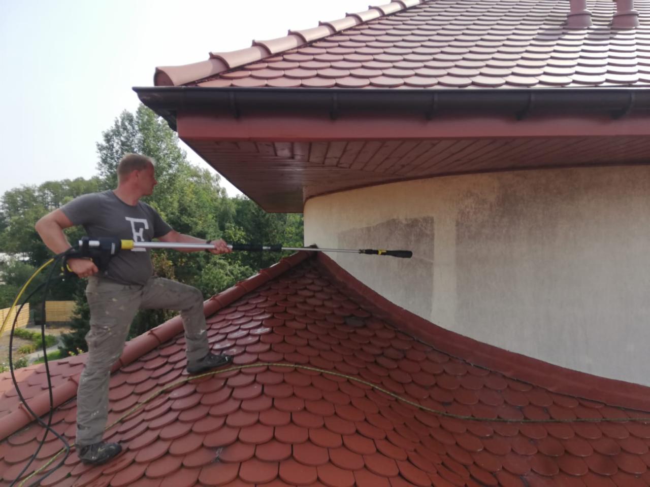 Mężczyzna w szarej koszulce i spodniach khaki, stojący na dachu krytym czerwoną dachówką, czyści elewację budynku za pomocą myjki ciśnieniowej z długą lancą. Widoczne ślady czyszczenia na jasnej...