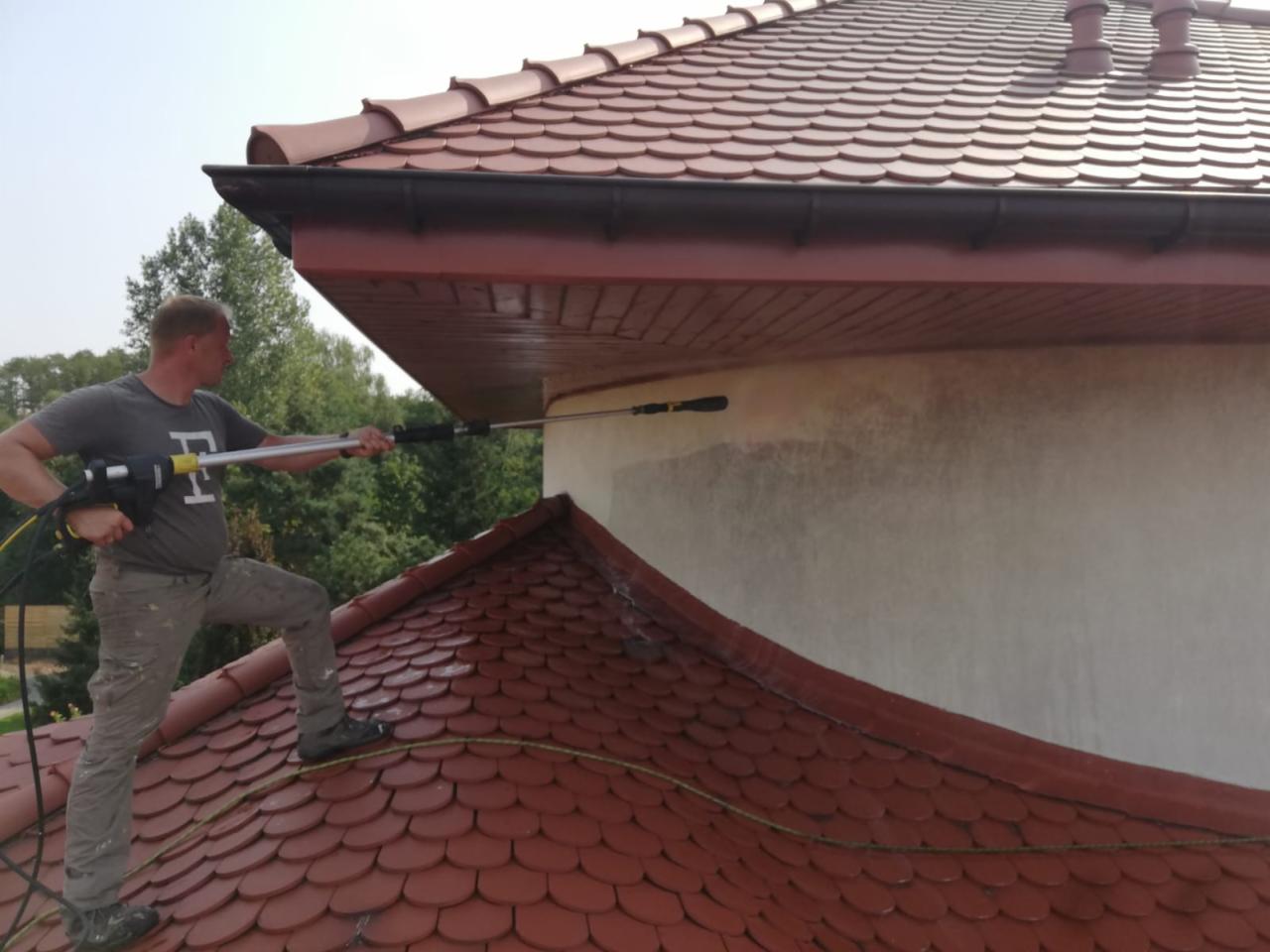 Mężczyzna w szarej koszulce i spodniach, stojąc na czerwonym dachu z dachówki, czyści jasną elewację domu za pomocą myjki ciśnieniowej z długą lancą. Widoczne ślady czyszczenia na elewacji.