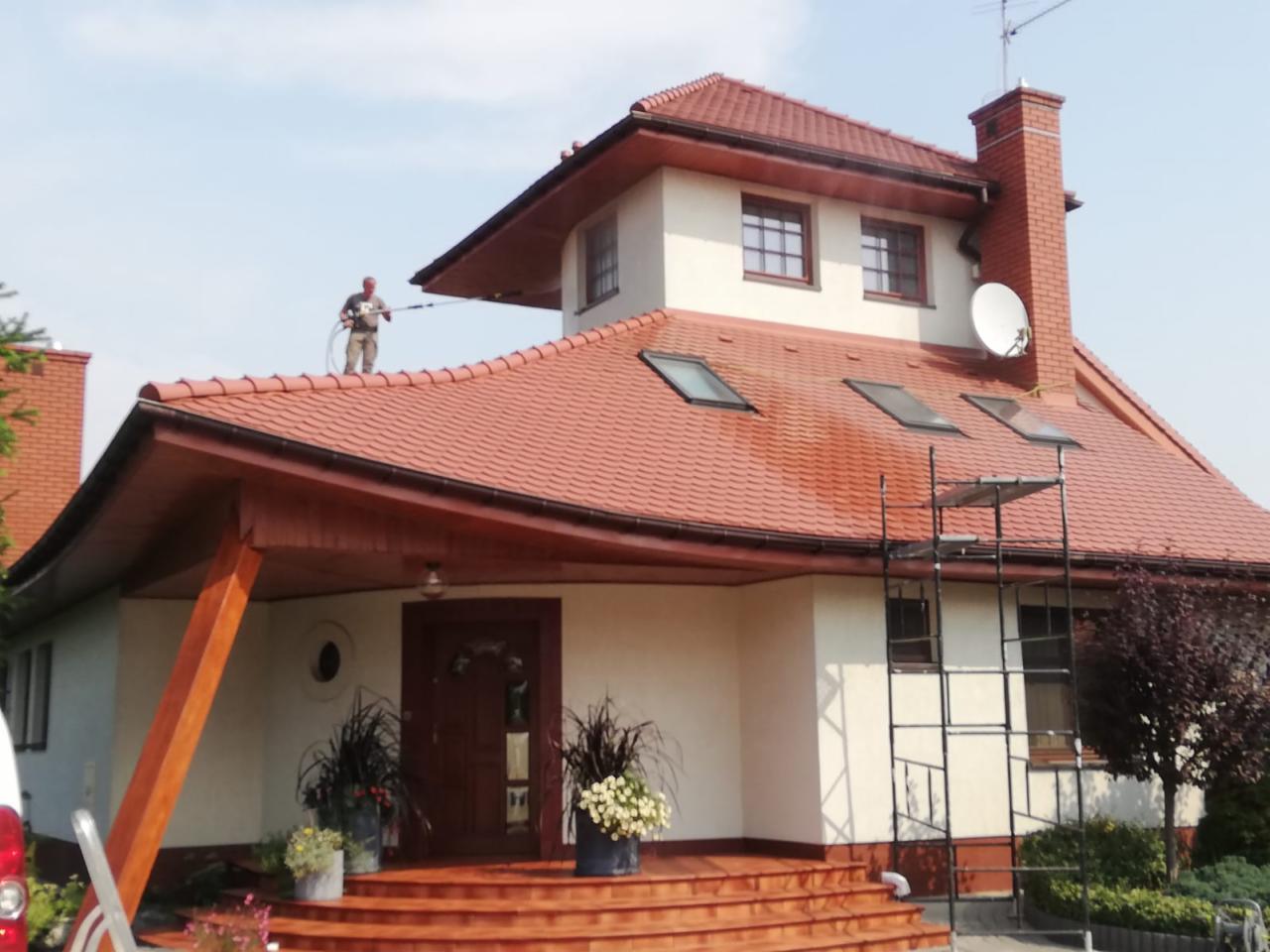 Pracownik w kombinezonie ciśnieniowo czyści czerwoną dachówkę domu jednorodzinnego z drabiny, widoczne zacieki i odbarwienia na dachu.