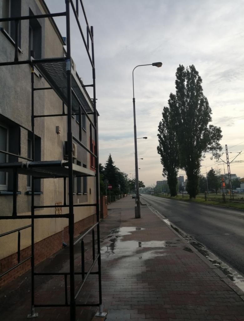 Metalowe rusztowanie ustawione wzdłuż ściany budynku z widocznymi oknami, chodnikiem i ulicą w tle, po deszczu.