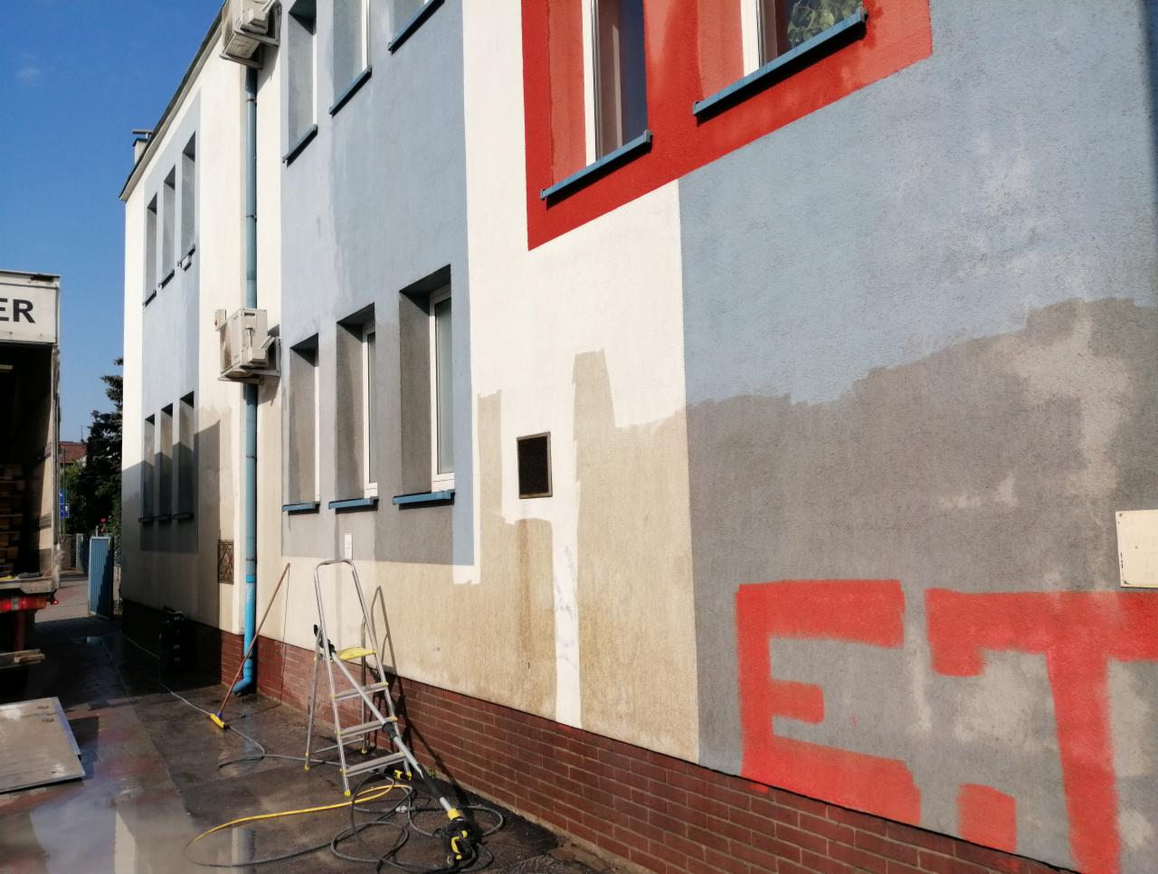 Budynek wielopiętrowy z fragmentami wyczyszczonej elewacji, widoczne ślady po usuniętych graffiti, drabina i sprzęt do czyszczenia pod ciśnieniem na chodniku.