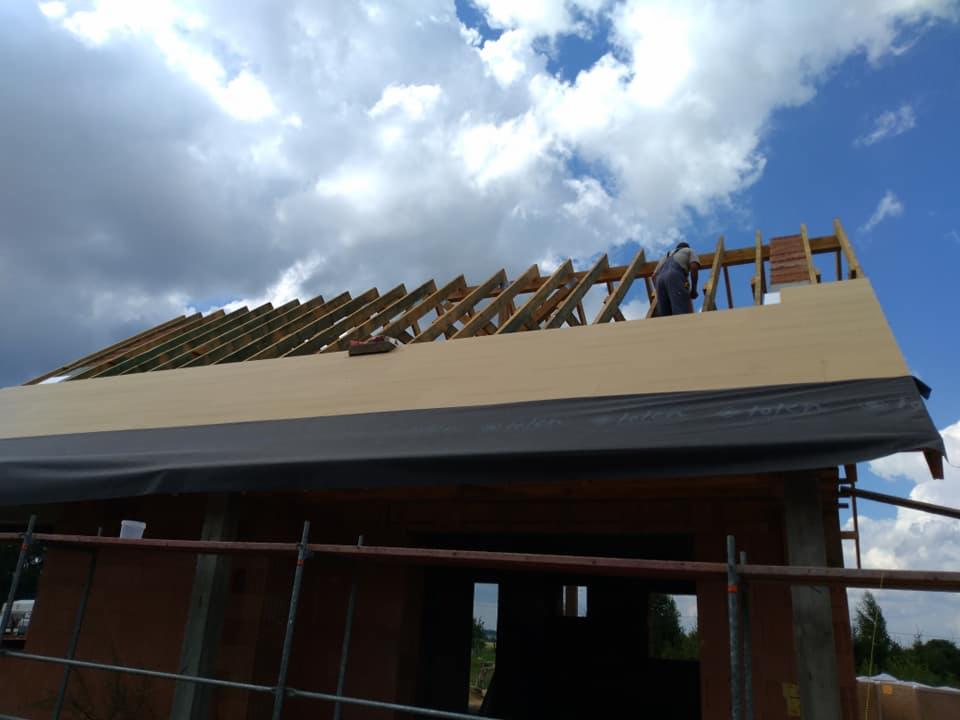 Wznoszenie dachu na surowym domu z cegły, widoczne krokwie, deski i czarna membrana dachowa, pracownik w kombinezonie na szczycie dachu, niebo z chmurami w tle.