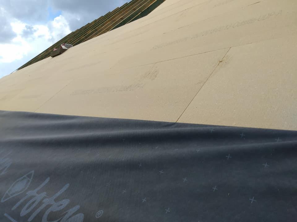 Ocieplanie dachu domu w trakcie budowy, widoczna membrana z nadrukiem, płyty izolacyjne i drewniana konstrukcja więźby dachowej.