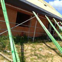 Surowy stan domu murowanego w trakcie budowy, widoczne zielone deski podtrzymujące konstrukcję, rusztowanie i fragment dachu z membraną, teren nierówny, trawiasty.