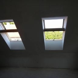 wieslaw - Dwa okna dachowe w szarej ścianie poddasza, jedno otwarte z widocznym drewnianym mechanizmem, drugie zamknięte z widokiem na zieleń na zewnątrz.