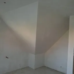 Poddasze po nałożeniu gładzi gipsowej, widoczne nierówności i przygotowanie do dalszych prac wykończeniowych, surowe wnętrze z widocznym punktem świetlnym na suficie.