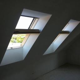 wieslaw - Dwa okna dachowe w nowo wykończonym poddaszu, widok na zewnątrz przez otwarte okno, ściany świeżo pomalowane na szaro, podłoga w trakcie przygotowania.