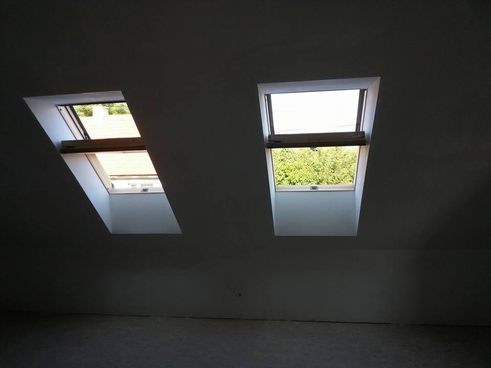 Dwa okna dachowe w szarej ścianie poddasza, jedno otwarte z widocznym drewnianym mechanizmem, drugie zamknięte z widokiem na zieleń na zewnątrz.