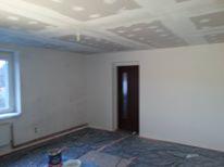 Pokój w trakcie prac wykończeniowych: częściowo zagruntowana ściana, sufit z widocznymi łączeniami płyt kartonowo-gipsowych przed wygładzeniem, podłoga zabezpieczona folią malarską, widoczne...