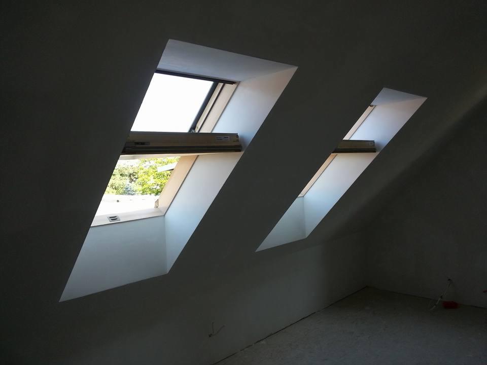 Dwa okna dachowe w nowo wykończonym poddaszu, widok na zewnątrz przez otwarte okno, ściany świeżo pomalowane na szaro, podłoga w trakcie przygotowania.