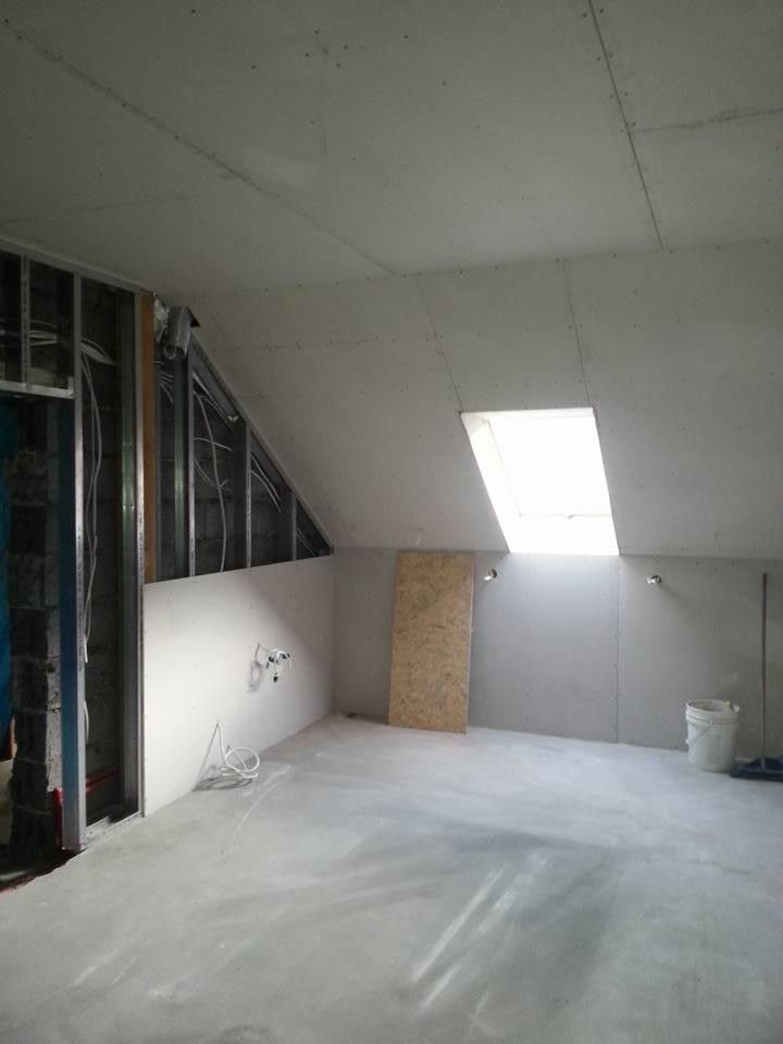 Poddasze w trakcie prac wykończeniowych: widoczne metalowe profile konstrukcyjne, częściowo otynkowane ściany, sufit z oknem dachowym, przygotowane do dalszych prac, z widocznymi przewodami...