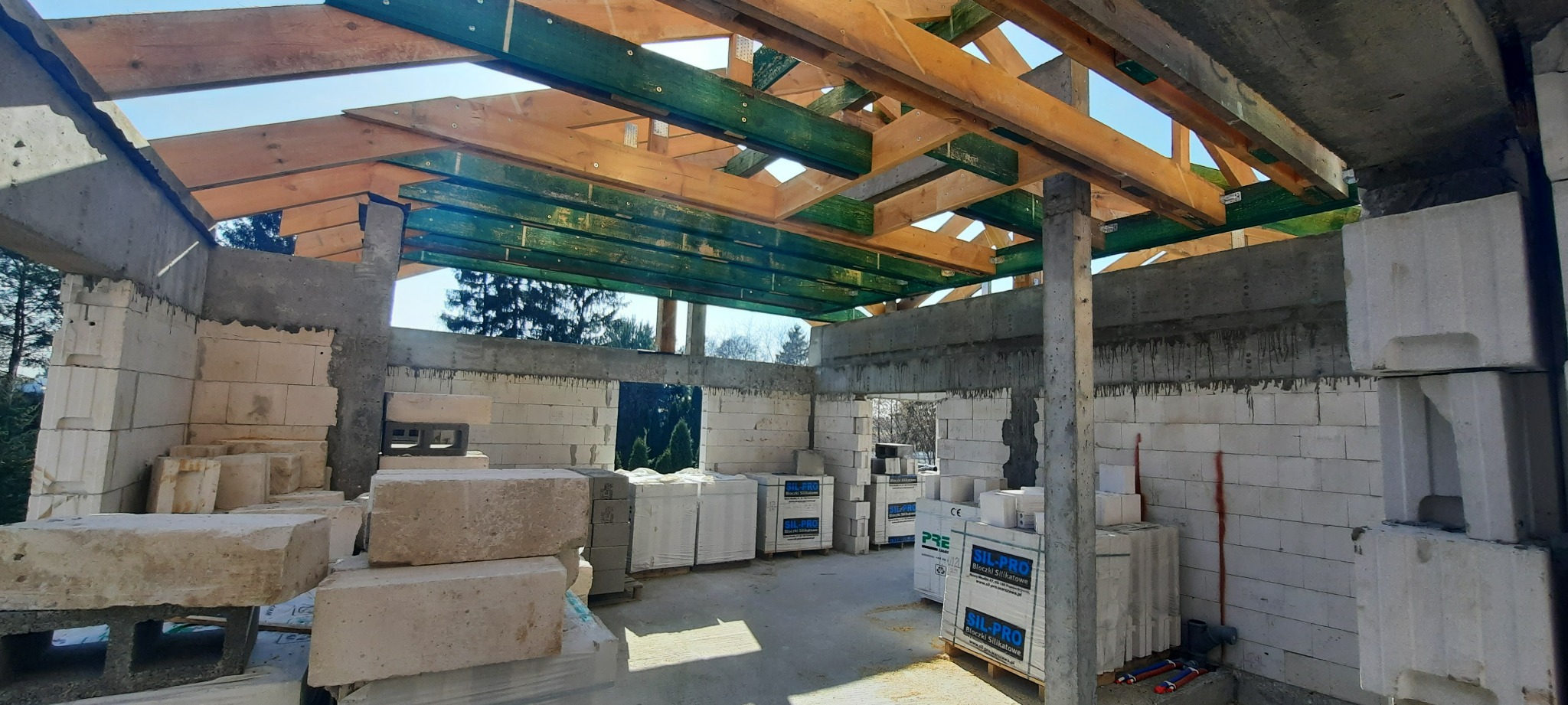 Wnętrze surowego budynku w trakcie budowy, widoczne ściany z białych bloczków silikatowych, drewniana konstrukcja dachu z elementami zabezpieczonymi na zielono, oraz palety z bloczkami SIL-PRO.