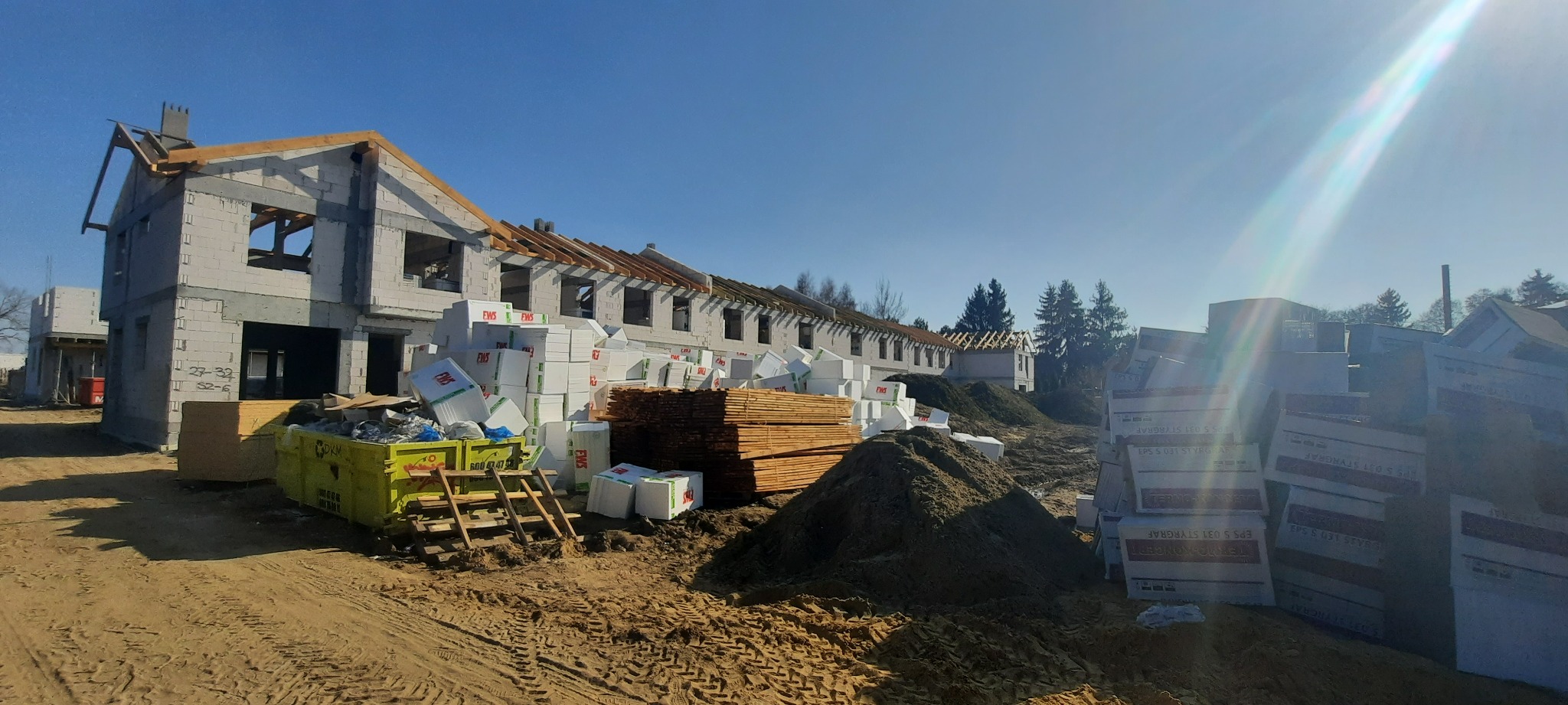 Wczesny etap budowy szeregu domów jednorodzinnych z widocznym szkieletem konstrukcyjnym, stertą styropianu i drewna budowlanego oraz kontenerem na odpady.