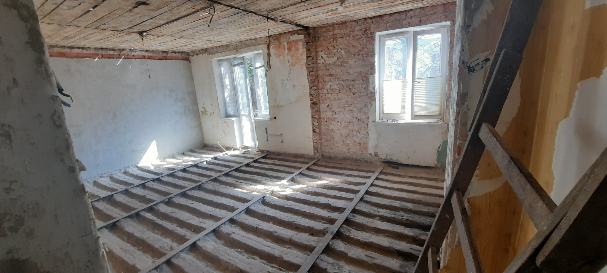 Pokój w trakcie remontu: odsłonięta cegła, częściowo otynkowane ściany, drewniany stelaż pod podłogę, widoczne pozostałości starej tapety i instalacji.