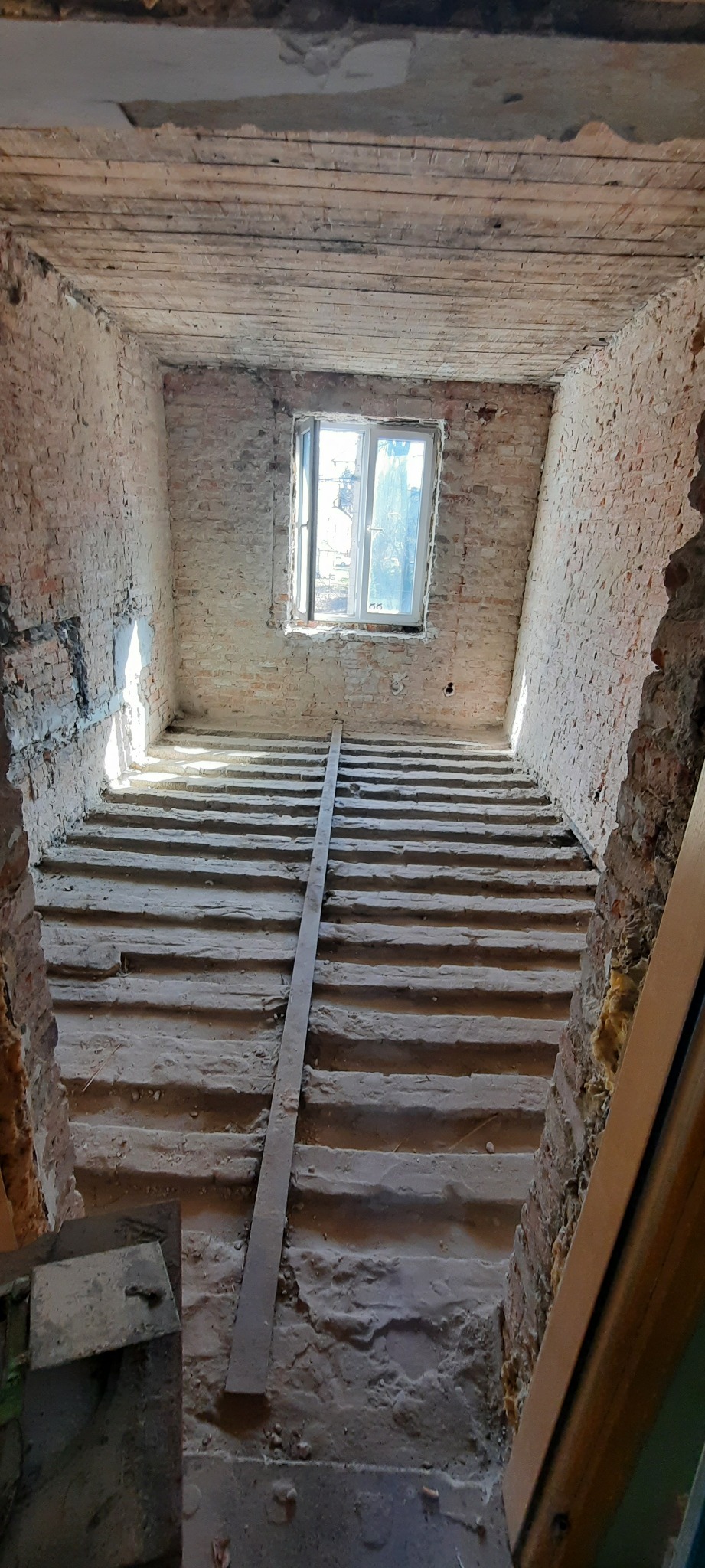 Wnętrze remontowanego budynku z odsłoniętą konstrukcją podłogi i ceglanymi ścianami, widoczne okno z widokiem na zewnątrz, surowy stan prac.