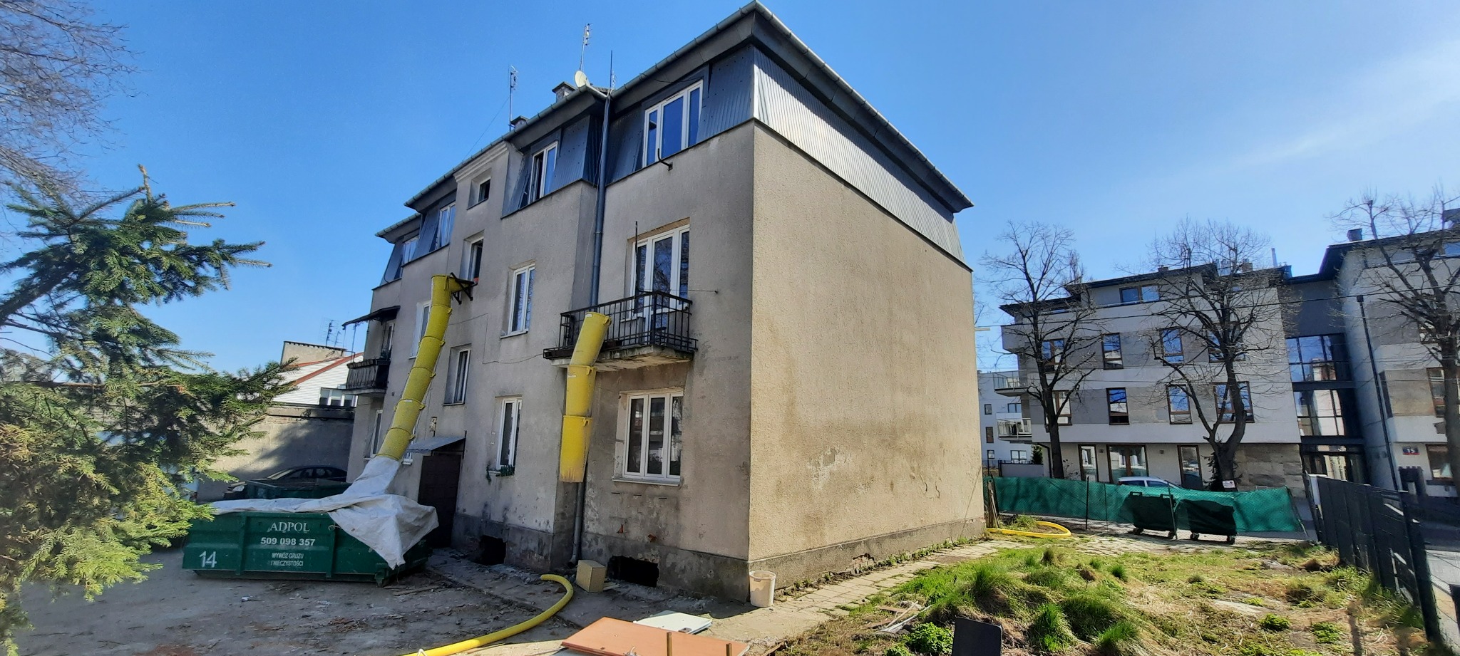 Blok mieszkalny w trakcie remontu, z żółtymi zsypami na gruz przymocowanymi do okien, kontener na odpady budowlane na trawniku.