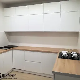 Nowoczesna kuchnia z białymi, lakierowanymi frontami szafek, drewnianym blatem i płytą grzewczą gazową oraz indukcyjną.