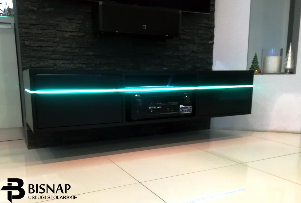 Czarna szafka RTV wisząca na ścianie z kamienia dekoracyjnego, podświetlona turkusowym światłem LED, z widocznym sprzętem audio w środkowej wnęce.