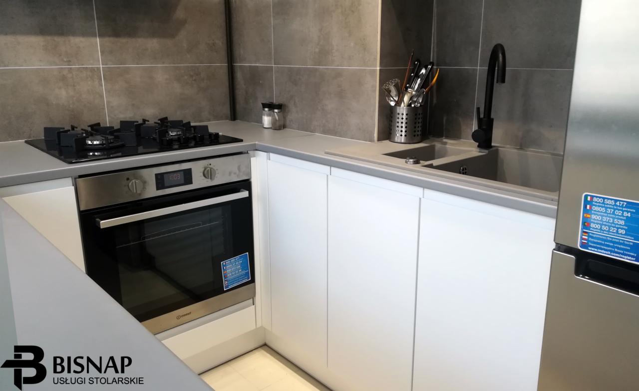 Nowoczesna kuchnia z białymi frontami szafek, szarym blatem i płytkami, czarną armaturą i sprzętem AGD ze stali nierdzewnej, widoczne logo firmy stolarskiej w lewym dolnym rogu.