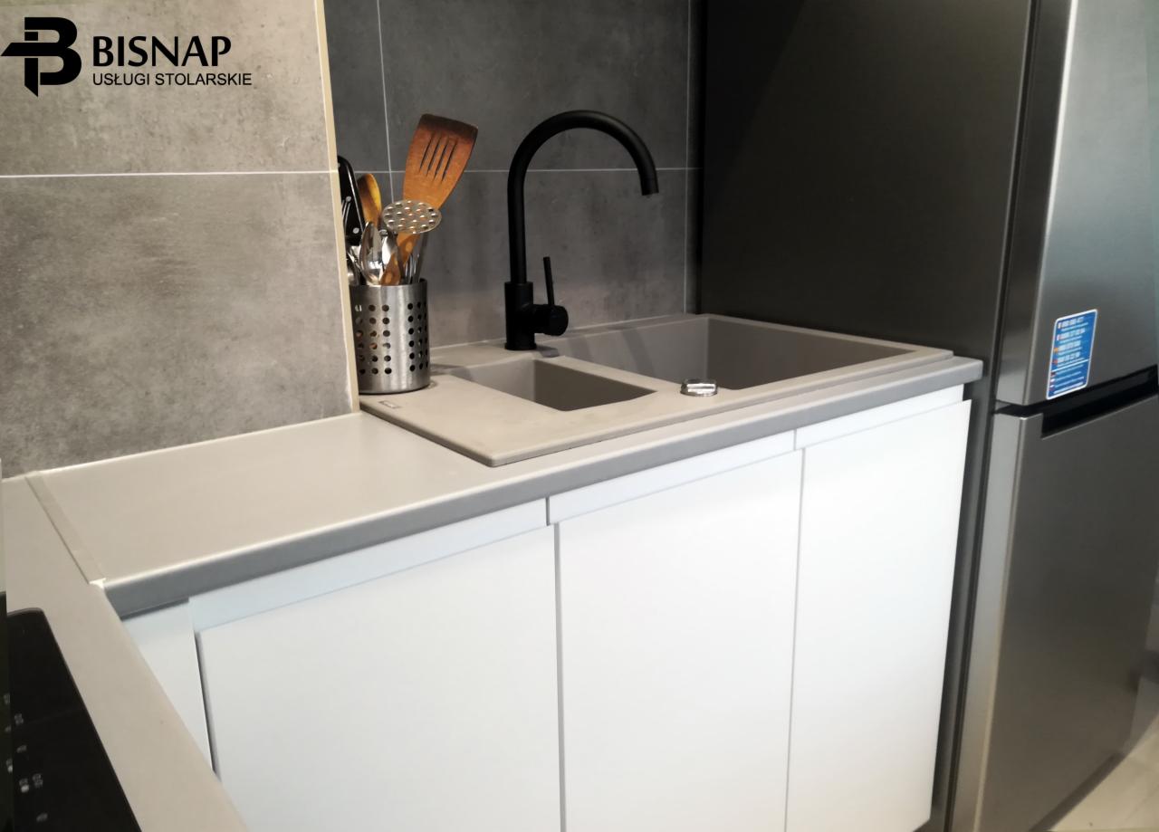 Szary blat kuchenny ze zlewem, czarną baterią i białymi szafkami pod spodem, widoczny obok lodówki, z logo firmy stolarskiej w lewym górnym rogu.