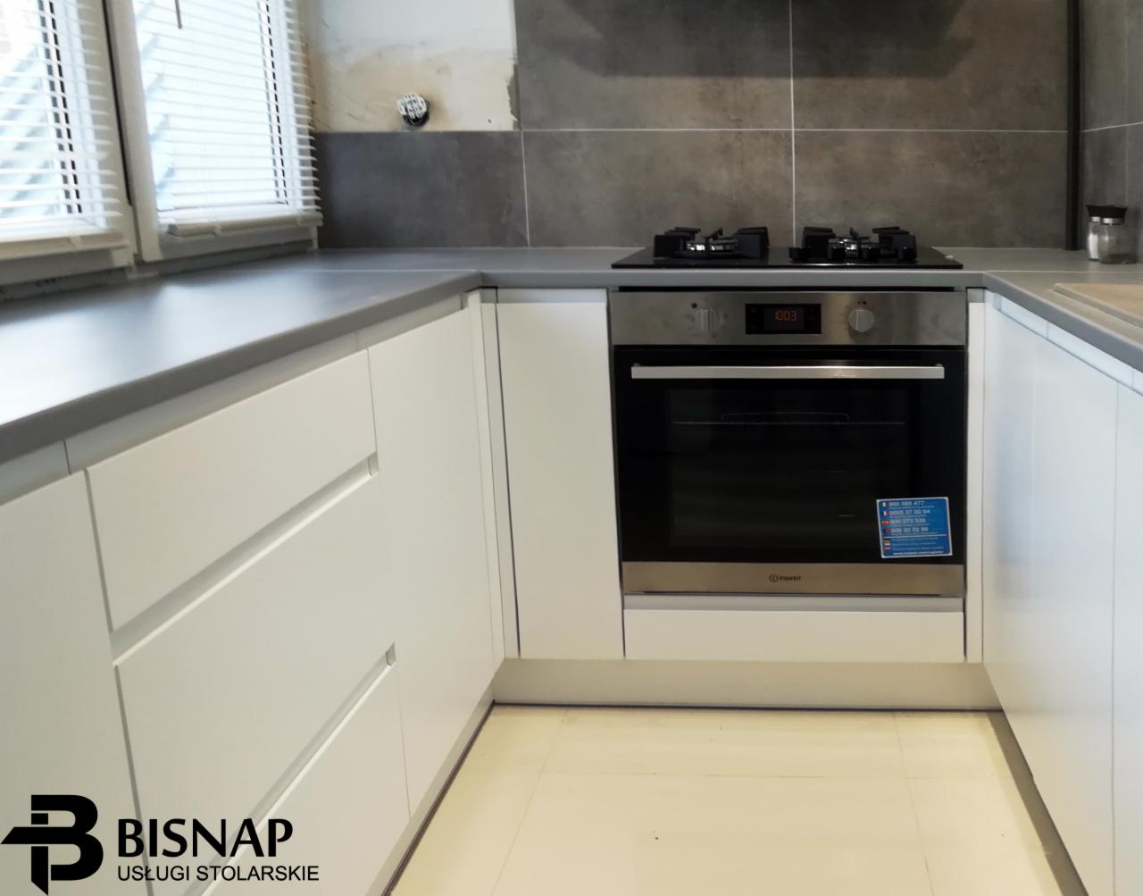 Nowoczesna kuchnia z białymi, minimalistycznymi szafkami, szarym blatem i wbudowanym piekarnikiem, częściowo wykończona szarą glazurą, widoczne logo firmy stolarskiej w rogu.