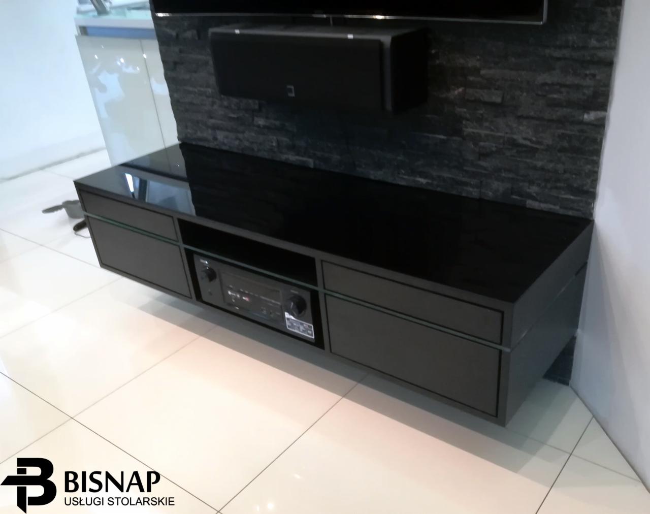 Czarna, lakierowana szafka RTV wisząca na ścianie z kamienia dekoracyjnego, z widocznym sprzętem audio w środkowej wnęce, oświetlona światłem odbitym od błyszczącej podłogi.