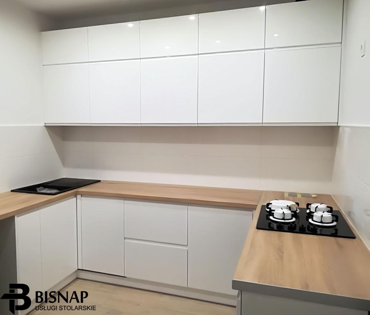 Nowoczesna kuchnia z białymi, lakierowanymi frontami szafek, drewnianym blatem i płytą grzewczą gazową oraz indukcyjną.