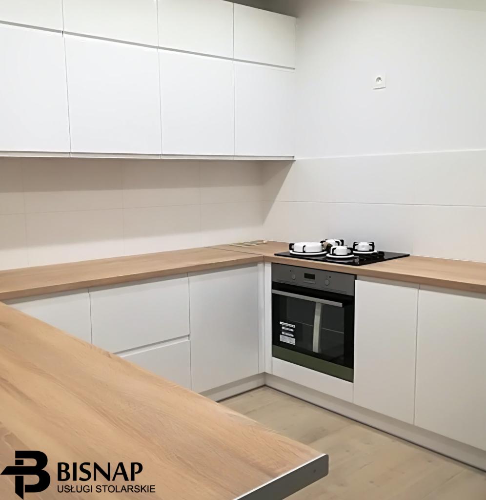Nowoczesna kuchnia w bieli z drewnianymi blatami, widoczny fragment wyspy kuchennej i logo firmy stolarskiej w rogu zdjęcia.