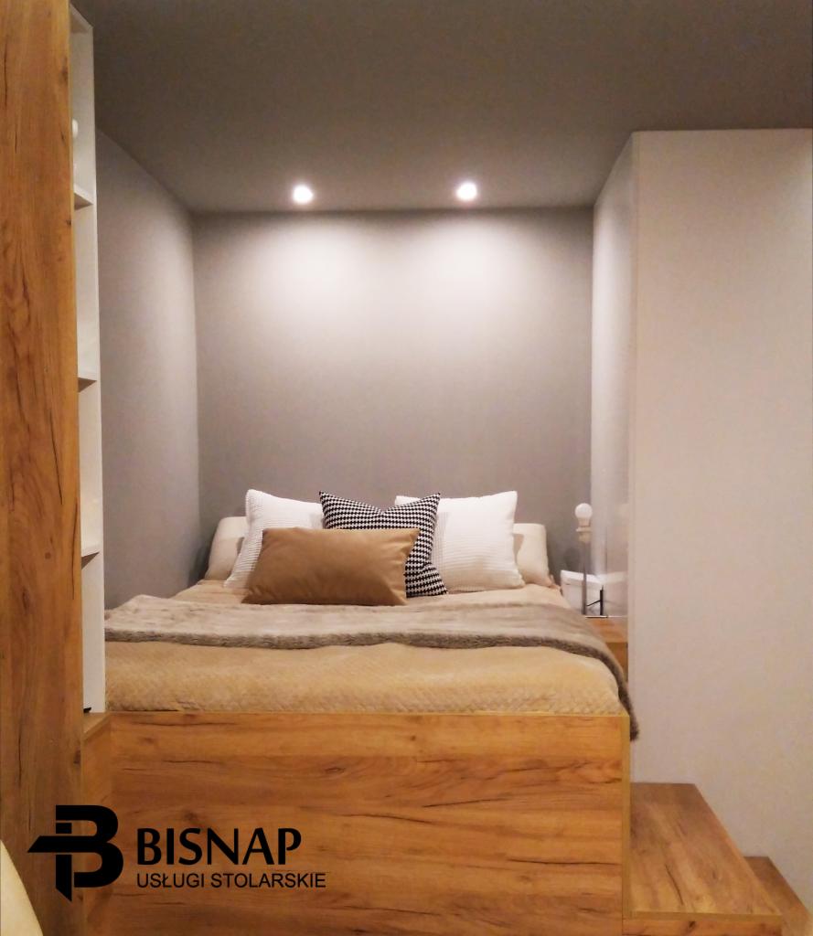 Drewniana platforma łóżka z widocznym fragmentem regału i schodkami, w małej sypialni z szarą ścianą i sufitowym oświetleniem punktowym, logo firmy stolarskiej w rogu.