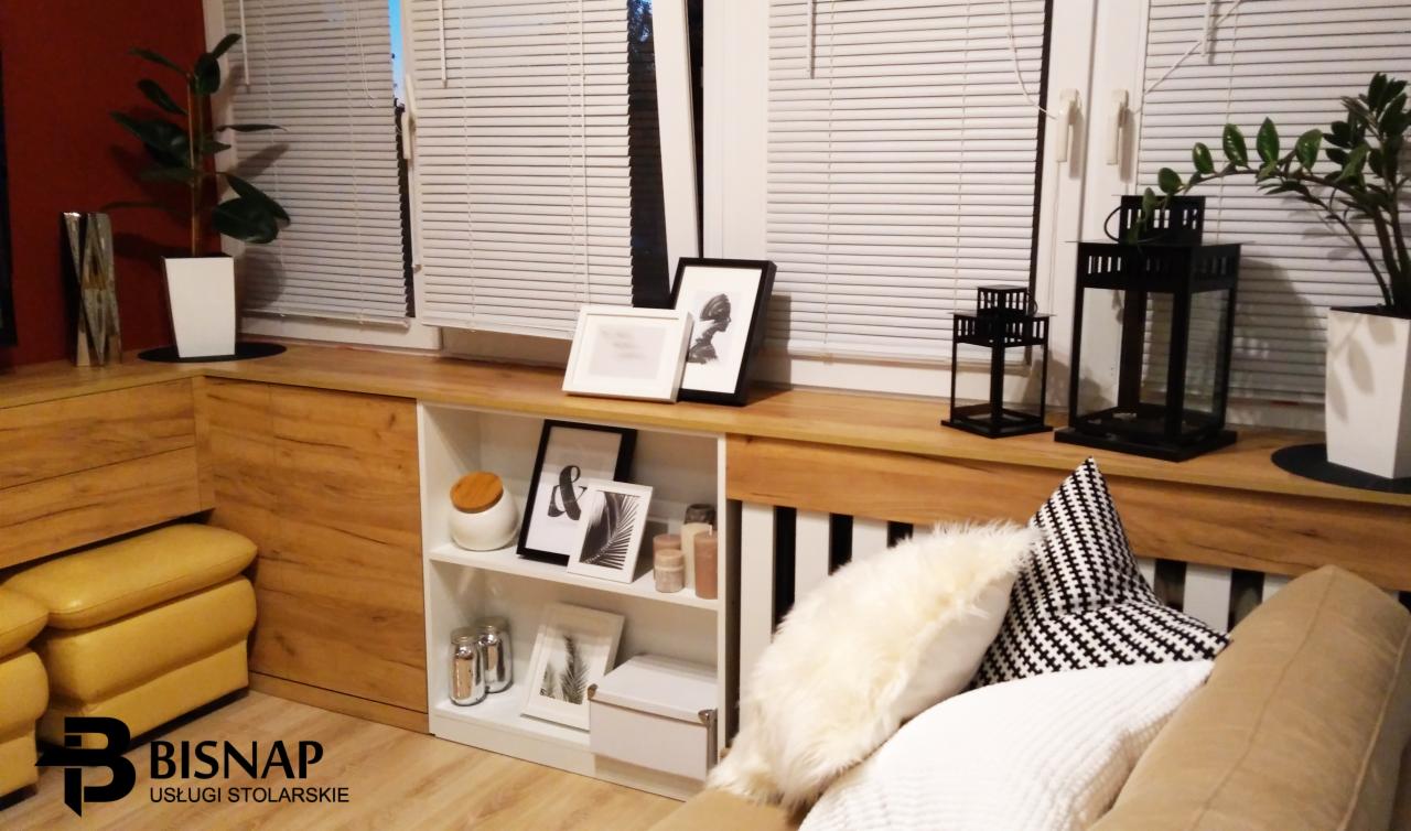 Aranżacja wnętrza z zabudową meblową pod oknem, drewnianym blatem i białymi półkami, dekoracje w postaci ramek, świeczników i roślin doniczkowych, widoczne logo firmy stolarskiej.
