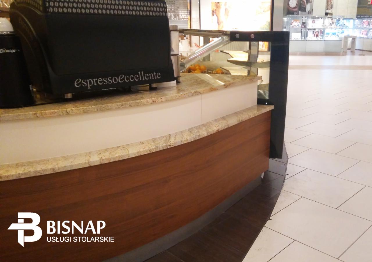 Kawiarniany lada z blatem z jasnego granitu i drewnianym frontem, ekspres do kawy z napisem 'Espressoeccellente', widoczne logo firmy stolarskiej w rogu zdjęcia.