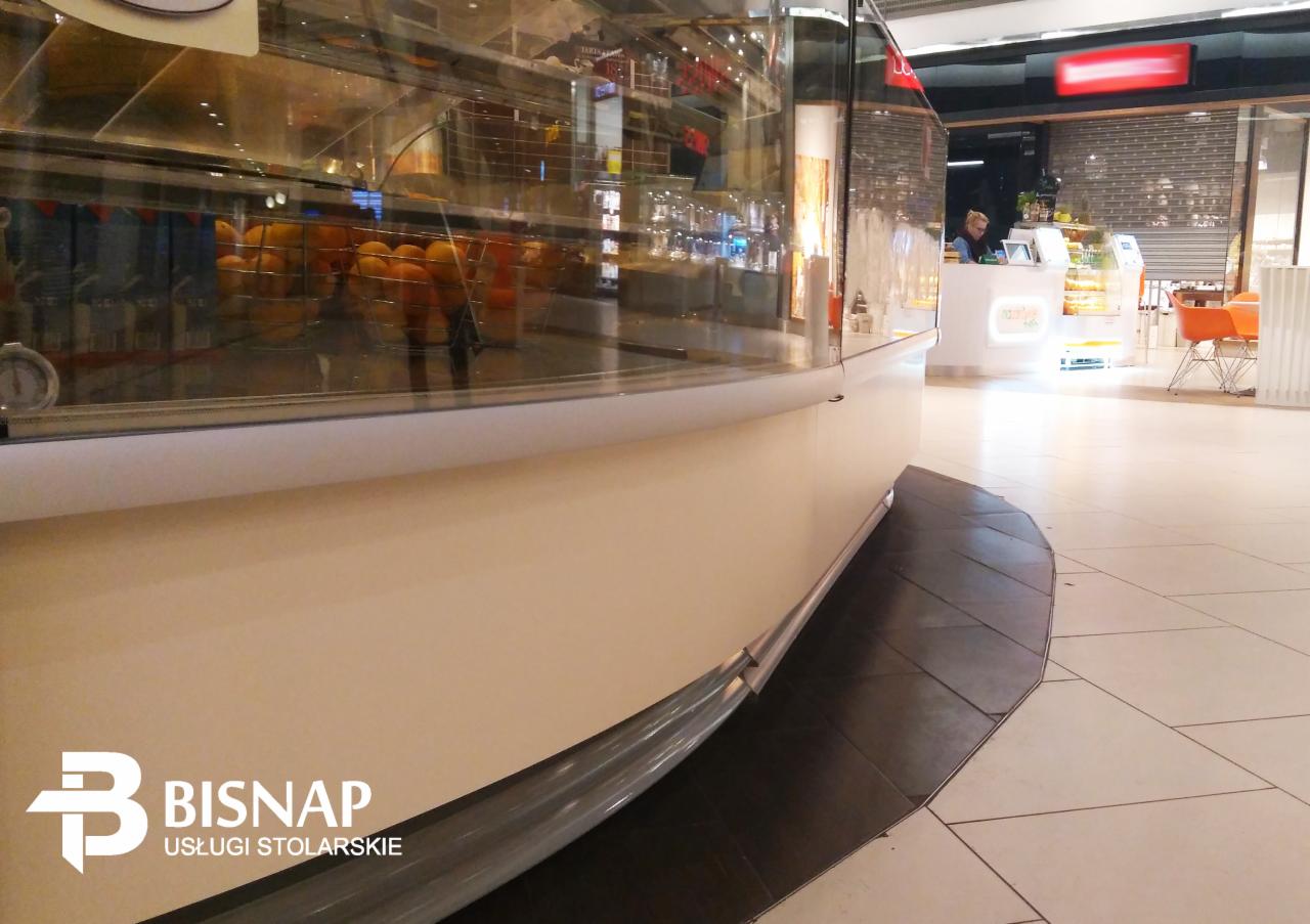 Stoisko handlowe z przeszkloną witryną, widoczne logo firmy Bisnap Usługi Stolarskie w lewym dolnym rogu, w tle inne stoisko z obsługą i pomarańczowe krzesła.